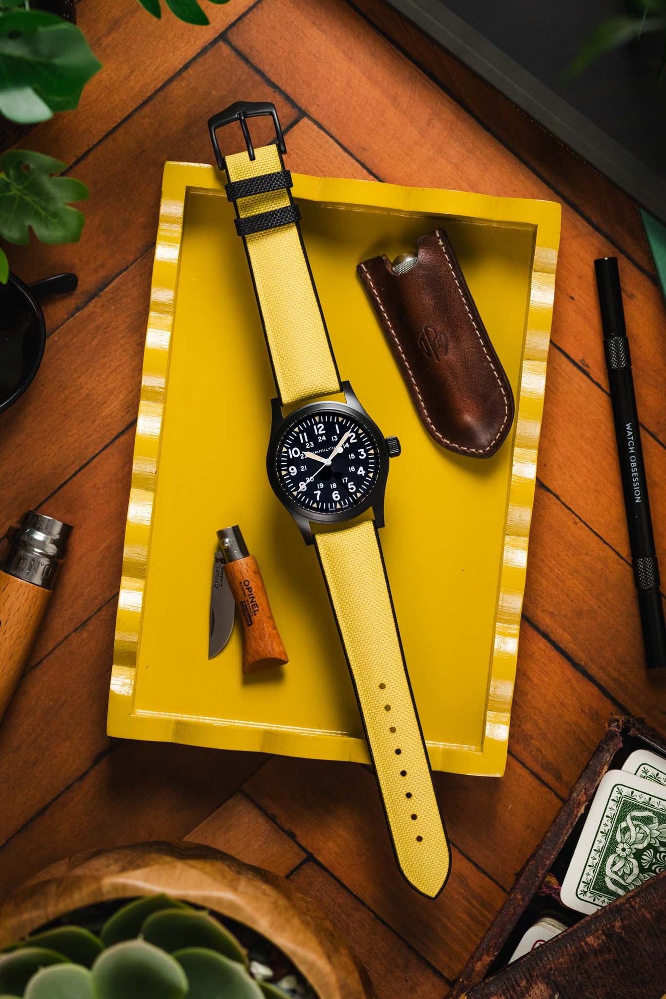 A black-cased Hamilton Khaki Field mechanical watch with white numerals and hands, paired with a bright yellow Hirsch Arne strap. It is placed on a yellow tray with vintage-style tins, playing cards, and a leather case.