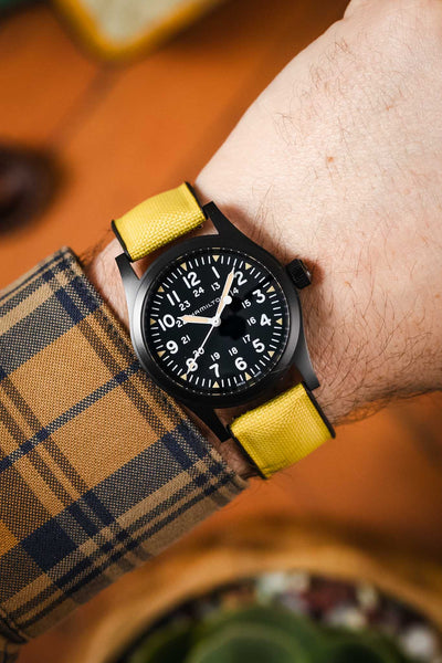 A close-up shot of a person's wrist wearing a black Hamilton Khaki Field Mechanical watch with a vibrant yellow Hirsch Arne Animal-Free Sailcloth Effect Performance strap. The watch features a black dial with white numerals and hands, including a 24-hour inner track. The person is wearing a blue and gold plaid shirt, and a blurred wooden surface is visible in the background.