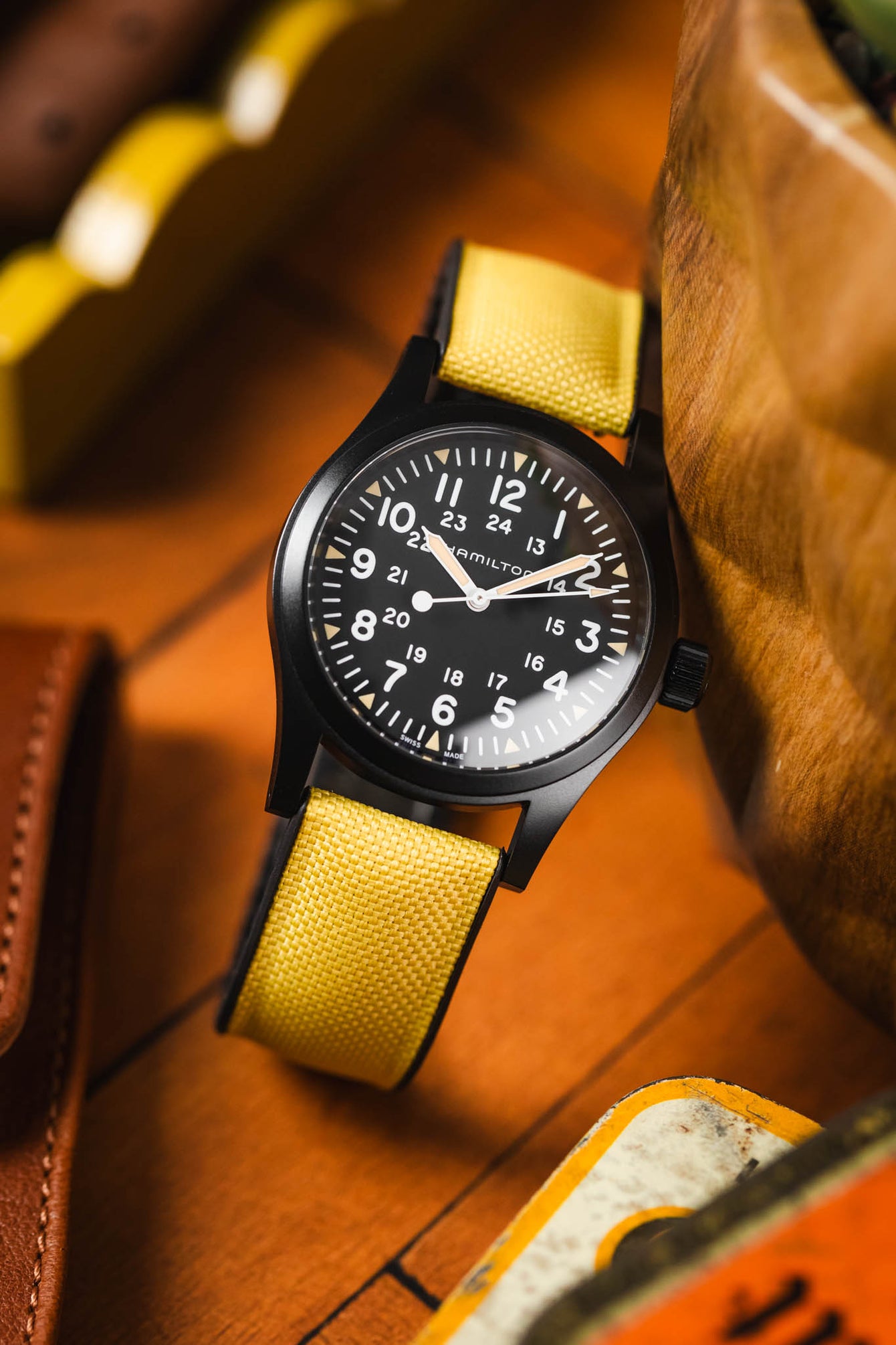 A black Hamilton Khaki Field Mechanical watch with a vibrant yellow Hirsch Arne Animal-Free Sailcloth Effect Performance strap is prominently displayed. The watch has a black dial with clear white numerals and hands, including a 24-hour inner track. The textured yellow strap is clearly visible. The watch rests on a wooden surface with blurred elements of a brown leather wallet and a vintage tin in the background, suggesting a casual, everyday setting.