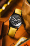 A black Hamilton Khaki Field Mechanical watch with a vibrant yellow Hirsch Arne Animal-Free Sailcloth Effect Performance strap is prominently displayed. The watch has a black dial with clear white numerals and hands, including a 24-hour inner track. The textured yellow strap is clearly visible. The watch rests on a wooden surface with blurred elements of a brown leather wallet and a vintage tin in the background, suggesting a casual, everyday setting.