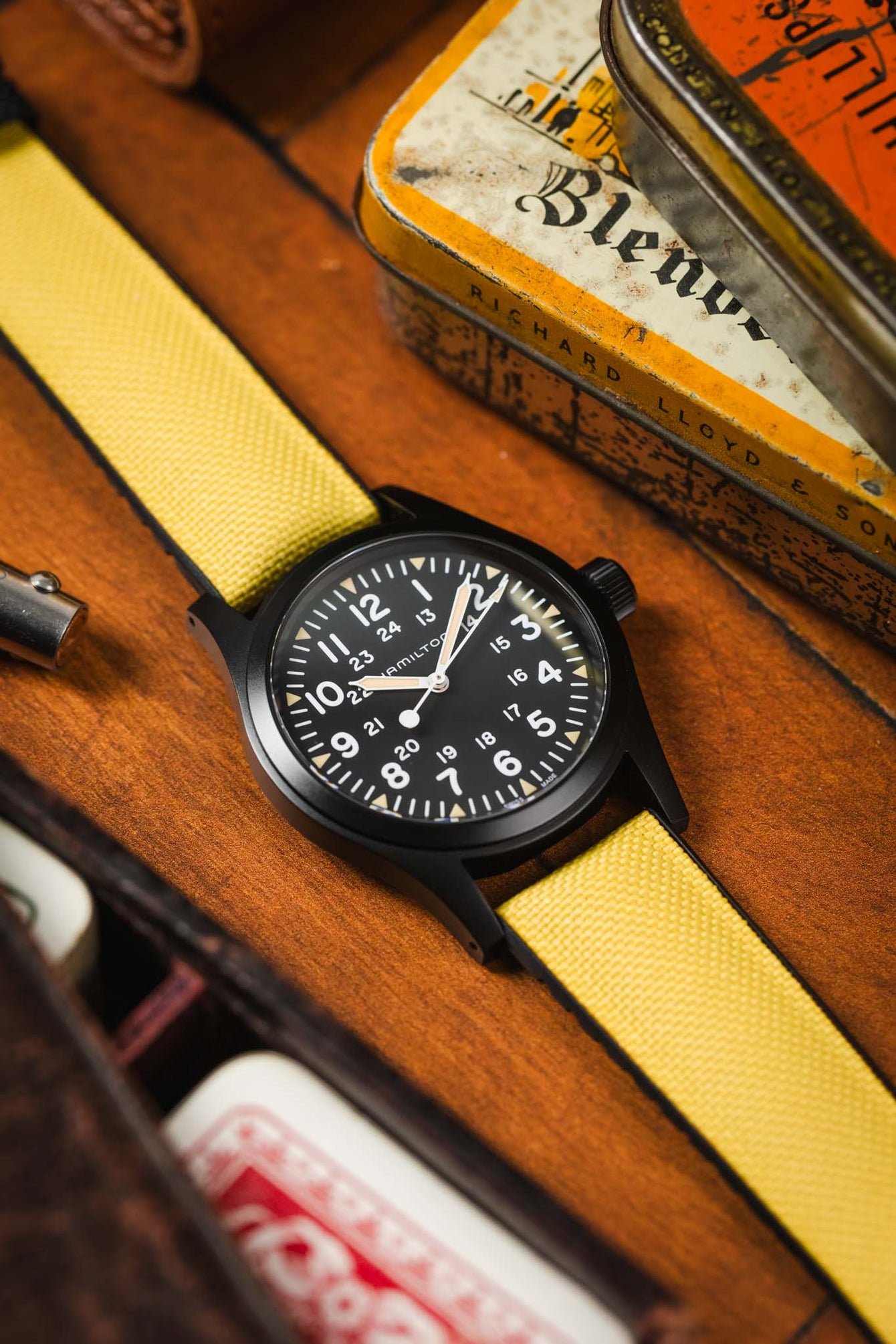 Close-up of a black-cased Hamilton Khaki Field mechanical watch featuring white numerals and hands, paired with a bright yellow Hirsch Arne strap. The watch rests on a textured wooden surface alongside vintage-style tins and playing cards.