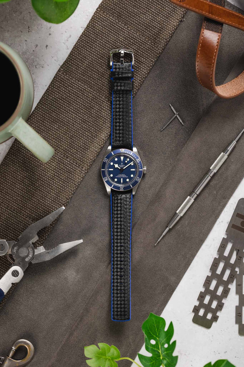 Flat lay of a stainless steel dive watch with a blue dial and bezel, fitted on a black perforated strap with blue edges, arranged on a textured surface. Surrounding items include a coffee cup, watchmaker’s screwdriver, metal caliper gauge, pliers, a brown leather strap, and green leaves, creating a workshop-style lifestyle setting against a mix of fabric and light grey background.