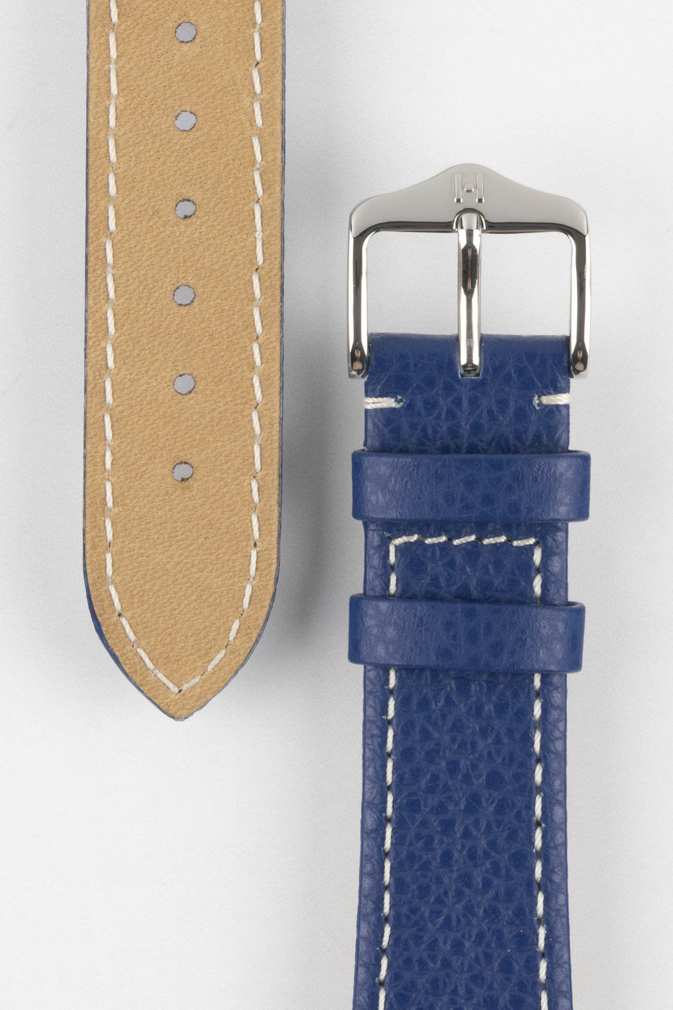 Close-up product image of a blue textured leather watch strap against a white background, showing the underside with multiple adjustment holes and beige lining on one section, and the other with a polished silver-tone pin buckle and two keepers, highlighting the white contrast stitching and fine grain texture.
