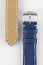 Close-up product image of a blue textured leather watch strap against a white background, showing the underside with multiple adjustment holes and beige lining on one section, and the other with a polished silver-tone pin buckle and two keepers, highlighting the white contrast stitching and fine grain texture.