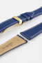 Close-up product image of a blue textured leather watch strap with white contrast stitching, displayed against a white background, featuring a polished gold-tone folding clasp and two keepers, alongside separate strap sections with exposed spring bars and a beige underside stamped with partial text “20 HIRSCH Bologna.”