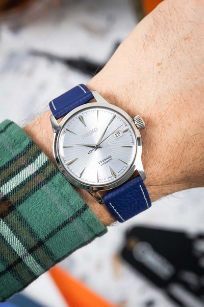 Close-up of a Seiko Presage watch with a textured white dial and date display, worn on a wrist with a blue leather strap featuring a grained finish and contrasting white stitching. The polished steel case and crown catch the light, while the strap wraps securely around the wrist with a snug fit. A green plaid sleeve sits just beneath the watch, and the softly blurred background with hints of tools and colour accents draws focus to the dial’s sunburst pattern and the strap’s rich texture.