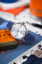 Close-up of a Seiko Presage watch with a textured white dial and date window at 3 o’clock, fitted on a blue leather strap with white stitching. The polished steel case rests on a vintage-style tin atop magazines, with a blurred background featuring bold orange and blue tones and surrounding desk items, creating a refined lifestyle scene that highlights the strap’s rich texture and colour.