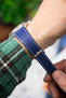 Close-up of a blue leather watch strap with a textured grain and contrasting white stitching, wrapped around a wrist. The strap is secured with a polished silver buckle and prong fastening through multiple adjustment holes, with one keeper loop holding the excess strap. A hand is visible adjusting the strap, while a green plaid sleeve sits just below the watch case. The softly blurred background and shallow depth of field draw attention to the strap’s rich colour, stitching, and fine leather detail.