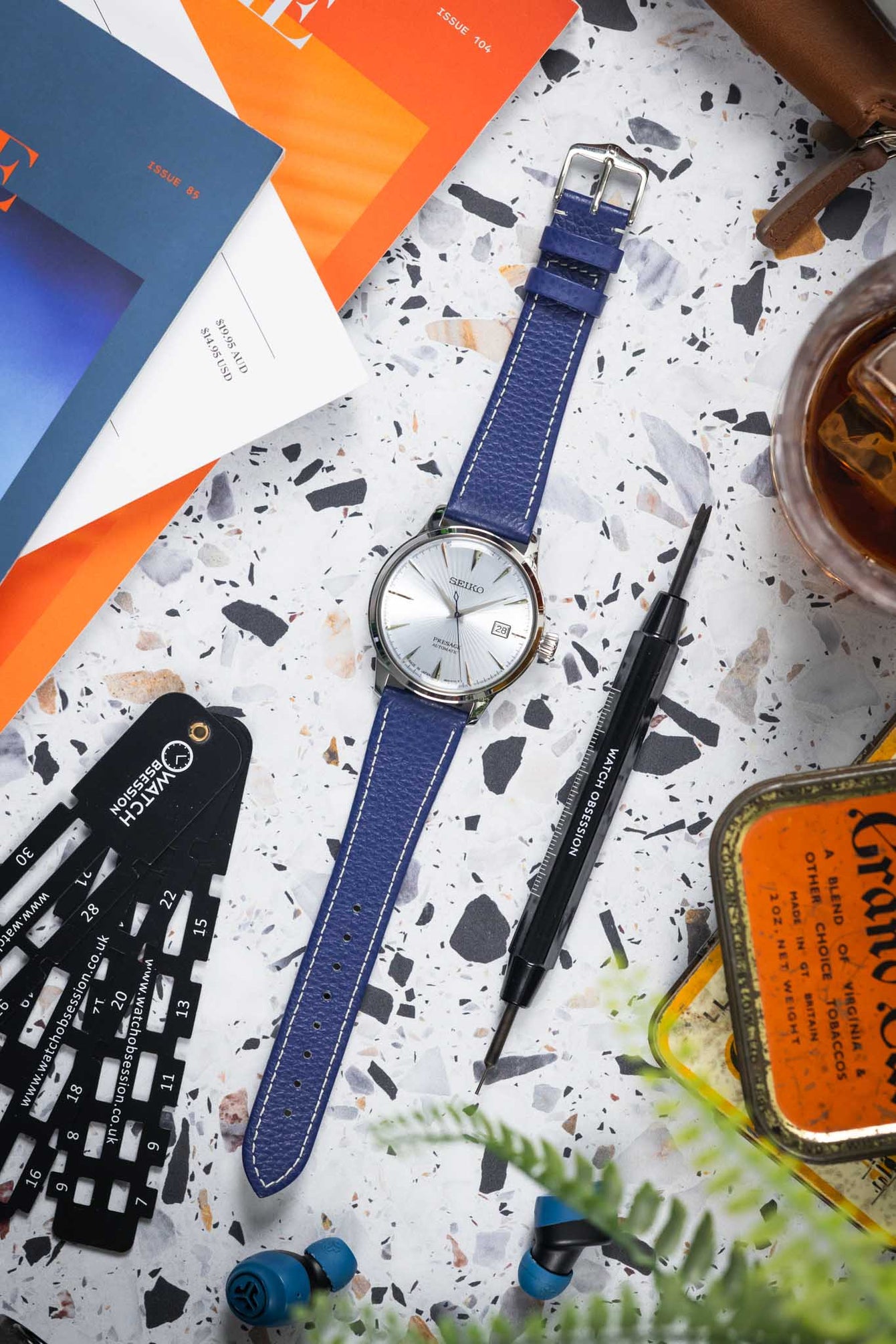 Flat-lay of a Seiko watch with a white dial on a blue leather strap with white stitching, arranged on a speckled surface. Surrounding objects include a watch sizing tool, a precision screwdriver, magazines, a glass with a drink, earbuds, and greenery, creating a lifestyle workspace scene with warm tones and soft lighting.
