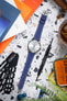 Flat-lay of a Seiko watch with a white dial on a blue leather strap with white stitching, arranged on a speckled surface. Surrounding objects include a watch sizing tool, a precision screwdriver, magazines, a glass with a drink, earbuds, and greenery, creating a lifestyle workspace scene with warm tones and soft lighting.