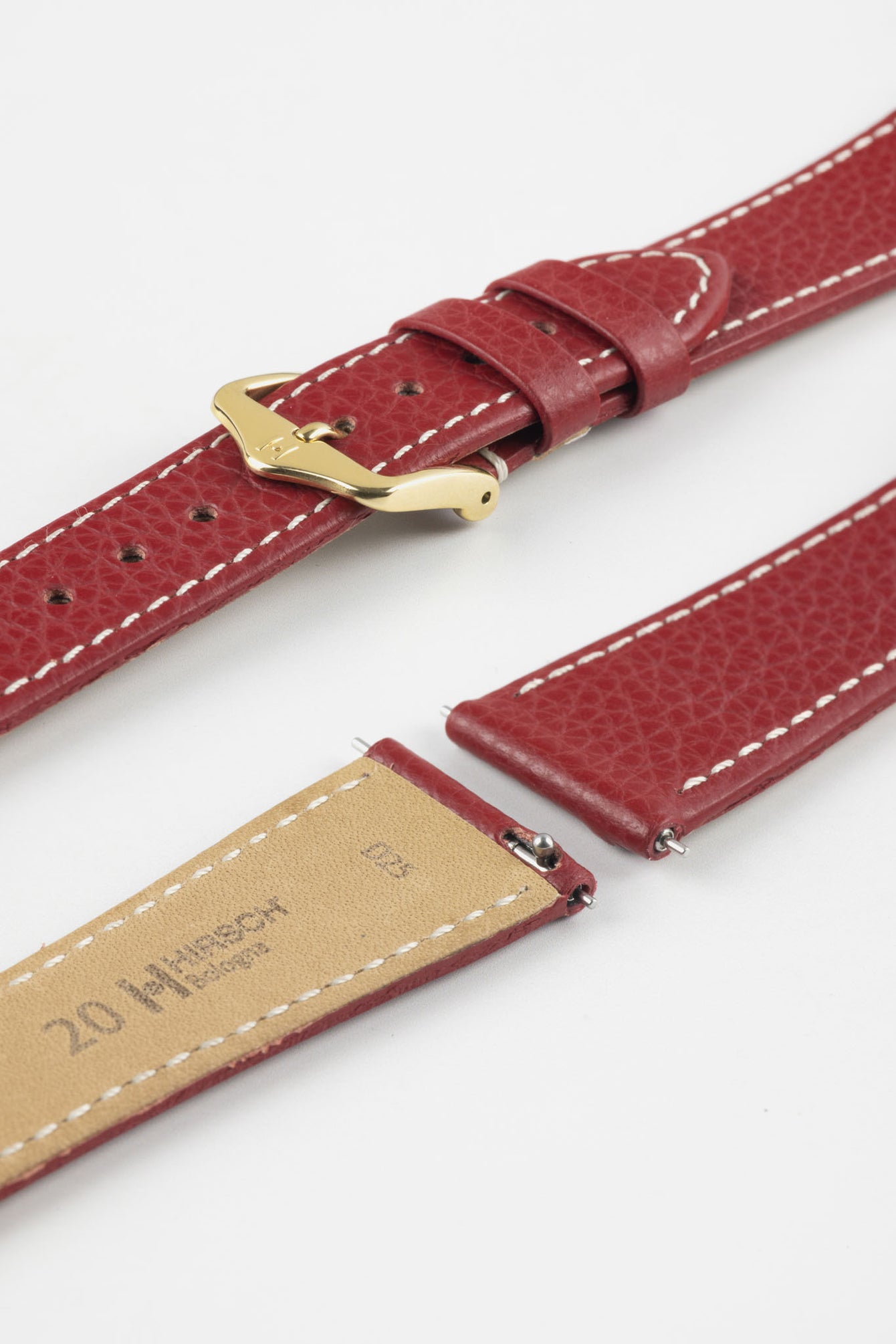Close-up product image of a red textured leather watch strap with white contrast stitching, displayed against a white background, featuring a polished gold-tone pin buckle with two keepers, alongside separate strap sections with exposed spring bars and a beige underside stamped with partial text “20 HIRSCH.”