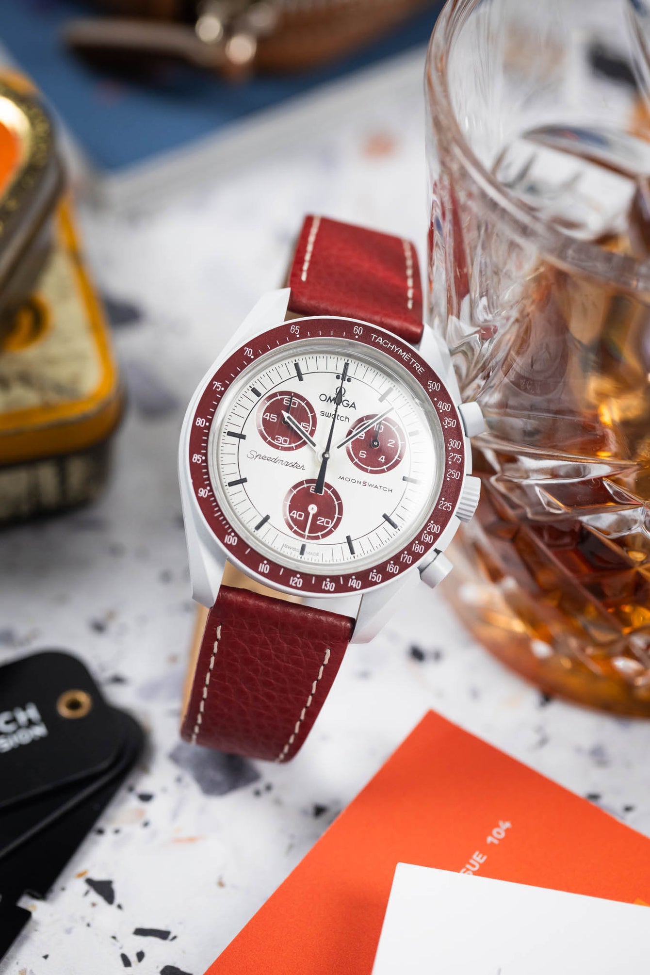 Omega x Swatch MoonSwatch with a white dial and burgundy bezel on a matching leather strap, styled beside a glass of whisky and accessories.