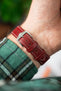 Close-up of a burgundy leather watch strap with a textured finish and contrasting white stitching, secured on a wrist. The strap features a polished silver buckle with a prong fastening through adjustment holes, along with two matching keeper loops. A green plaid sleeve is visible near the wrist, and the softly blurred background highlights the strap’s rich colour and grain.