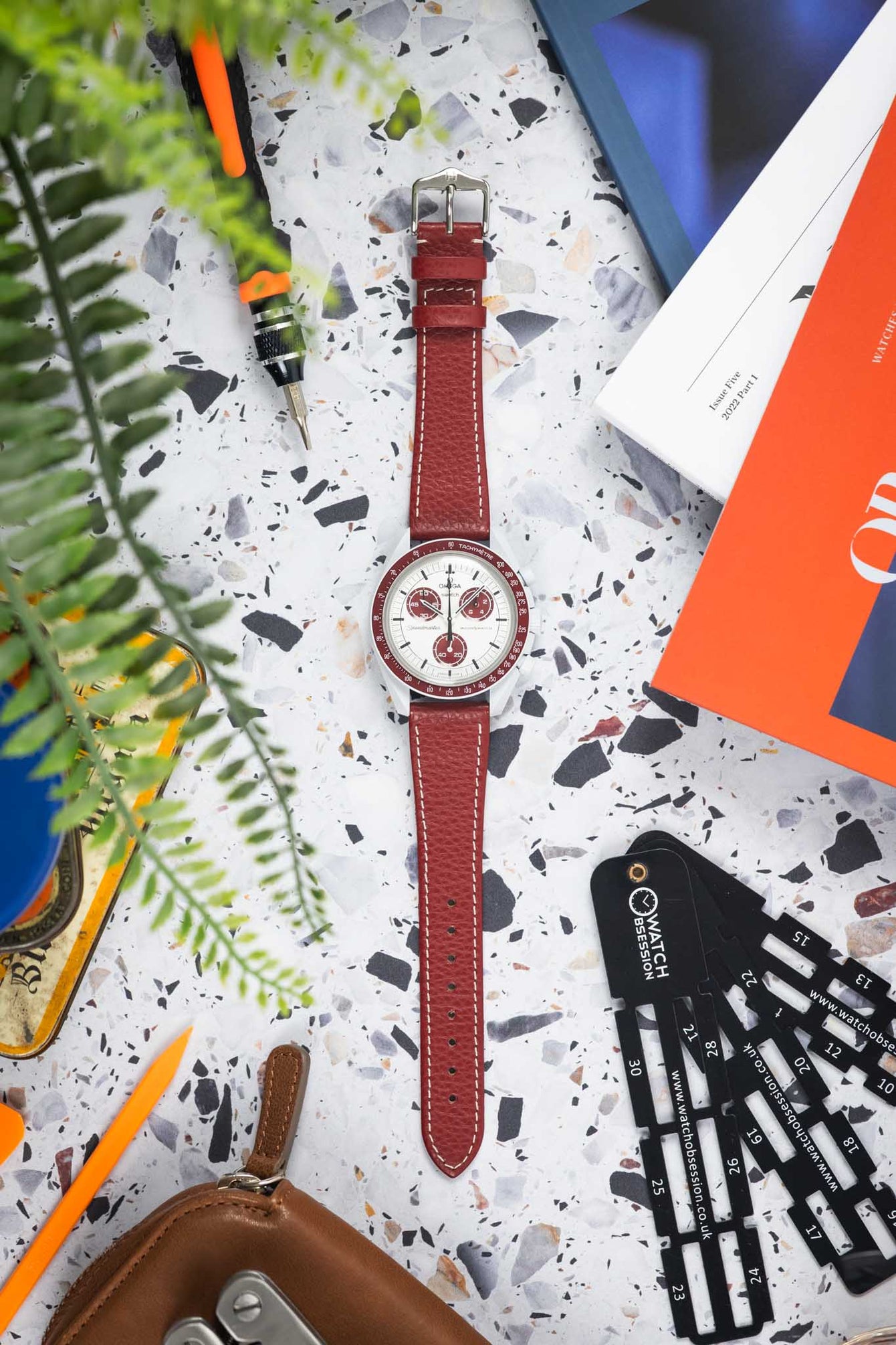 Flat-lay of an Omega x Swatch MoonSwatch with burgundy strap on a speckled surface, surrounded by a pen nib, book, greenery, and watch tools.