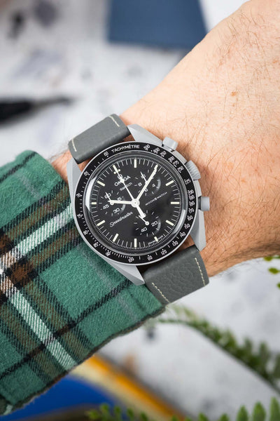 Close-up of a black-dial chronograph wristwatch with white markers and subdials, worn on a wrist with a grey leather strap featuring white stitching. The watch has a tachymeter bezel and is paired with a green plaid sleeve, set against a softly blurred background with hints of indoor objects and greenery.
