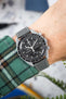 Close-up of a black-dial chronograph wristwatch with white markers and subdials, worn on a wrist with a grey leather strap featuring white stitching. The watch has a tachymeter bezel and is paired with a green plaid sleeve, set against a softly blurred background with hints of indoor objects and greenery.