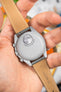 Back of a grey Omega x Swatch MoonSwatch watch held in a hand, showing a moon graphic and “MISSION TO THE MOON” text. The grey leather strap with white stitching has a tan underside stamped “HIRSCH Bologna” and size markings.