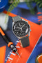 Close-up of a black Omega x Swatch Speedmaster MoonSwatch wristwatch with a grey leather strap, angled diagonally over a bright orange and blue background. The watch features a tachymeter bezel, chronograph subdials, and white markers on a black dial. A fountain pen nib and a second strap are partially visible in the foreground, while soft green leaves appear blurred in the background.