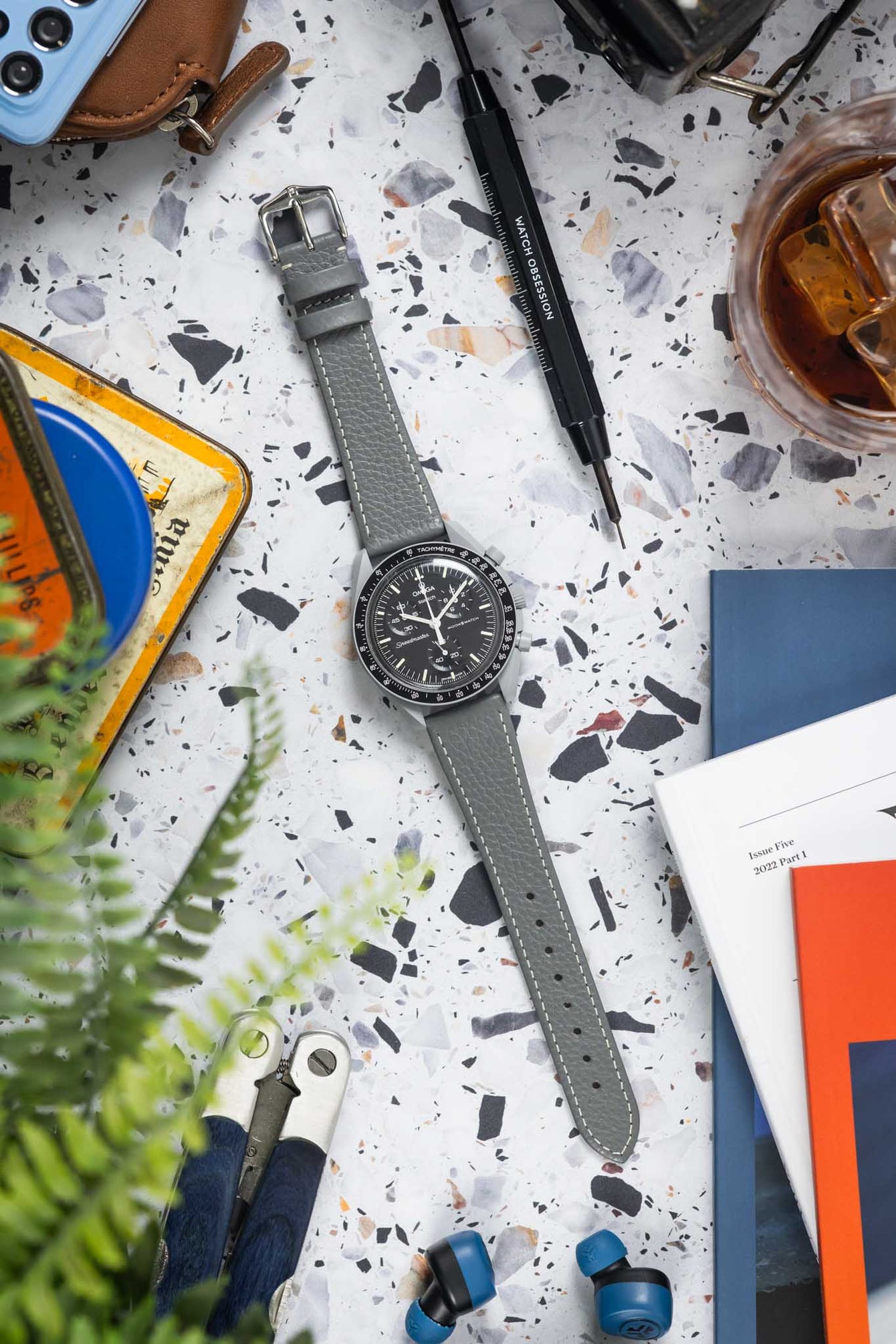 Top-down view of a black-dial chronograph wristwatch with white markers and subdials, attached to a grey leather strap with white stitching, laid flat on a speckled surface. The watch is surrounded by everyday items including a watch tool labeled “WATCH OBSESSION,” a glass of iced drink, a brown leather pouch, blue wireless earbuds, colorful printed cards, a tool set, and partially blurred green plant leaves in the foreground.