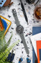 Top-down view of a black-dial chronograph wristwatch with white markers and subdials, attached to a grey leather strap with white stitching, laid flat on a speckled surface. The watch is surrounded by everyday items including a watch tool labeled “WATCH OBSESSION,” a glass of iced drink, a brown leather pouch, blue wireless earbuds, colorful printed cards, a tool set, and partially blurred green plant leaves in the foreground.