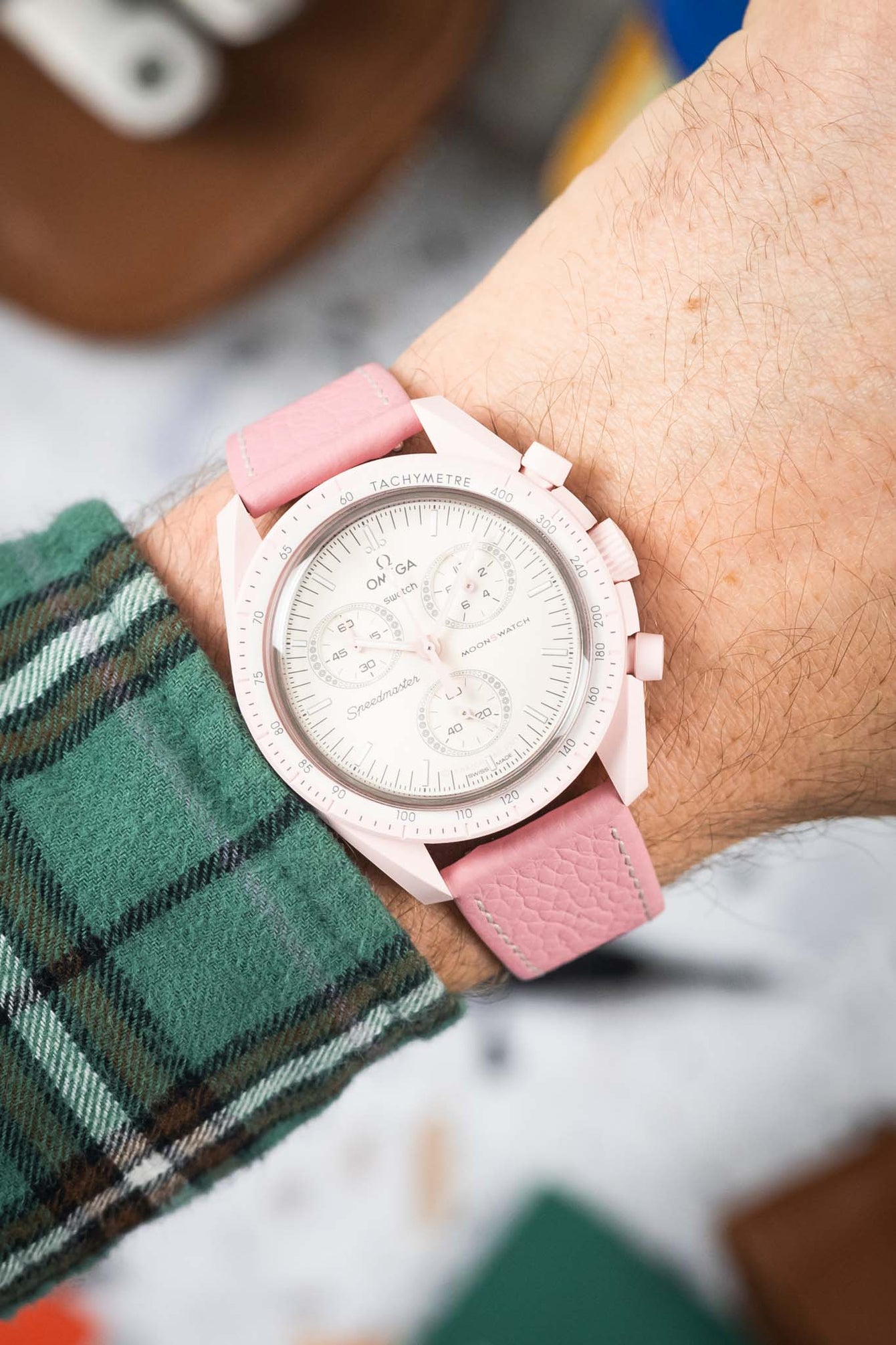Close-up of a pink chronograph wristwatch with a white dial and subdials, worn on a wrist with a pink leather strap and white stitching; a green plaid sleeve is visible, with a softly blurred background.