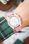 Close-up of a pink chronograph wristwatch with a white dial and subdials, worn on a wrist with a pink leather strap and white stitching; a green plaid sleeve is visible, with a softly blurred background.
