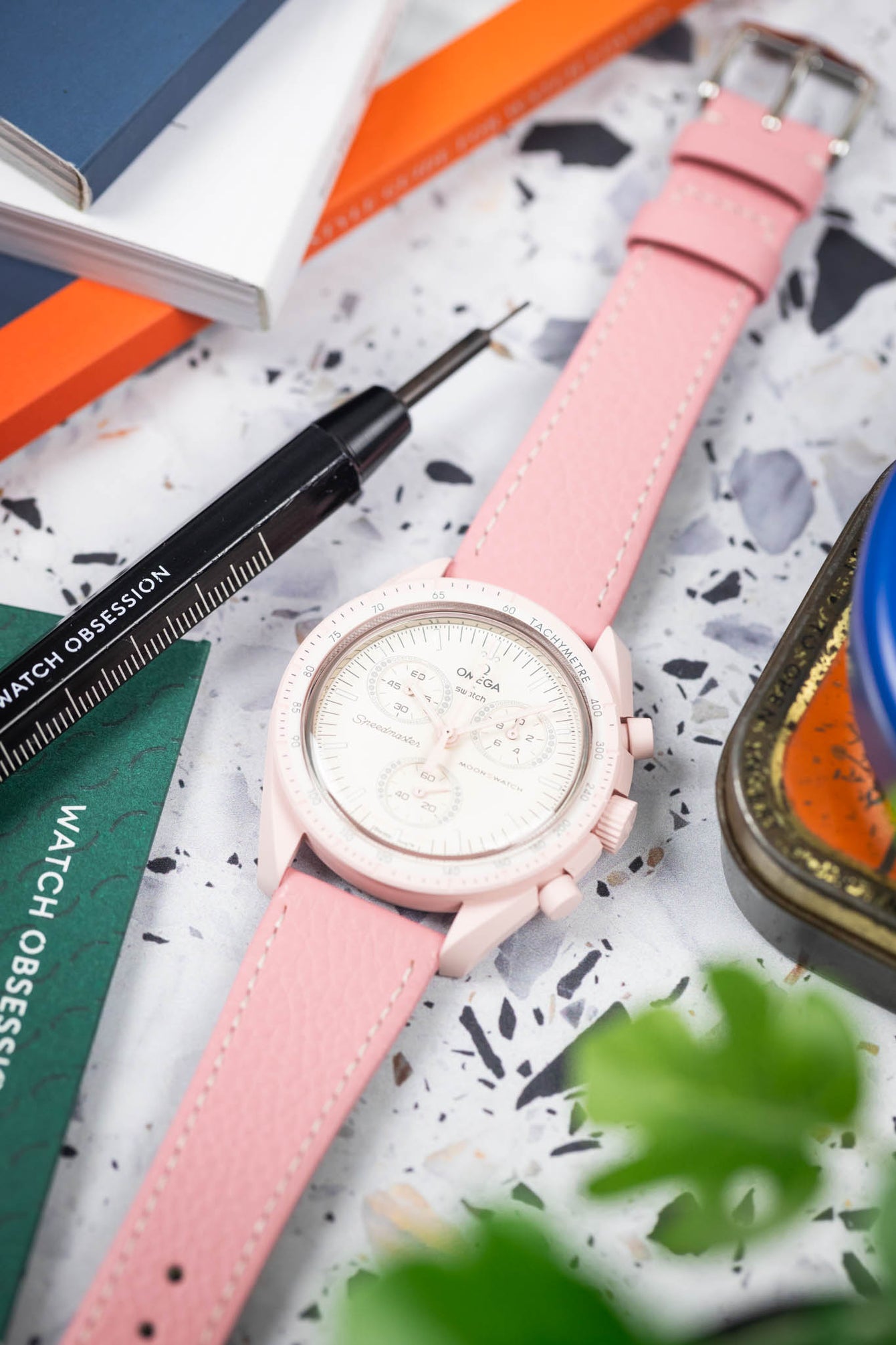 Close-up of a pink chronograph wristwatch with a white dial and subdials, paired with a pink leather strap and white stitching, placed on a speckled surface alongside a precision watch tool, colorful cards, and partially blurred greenery in the foreground.