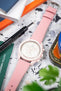 Close-up of a pink chronograph wristwatch with a white dial and subdials, paired with a pink leather strap and white stitching, placed on a speckled surface alongside a precision watch tool, colorful cards, and partially blurred greenery in the foreground.