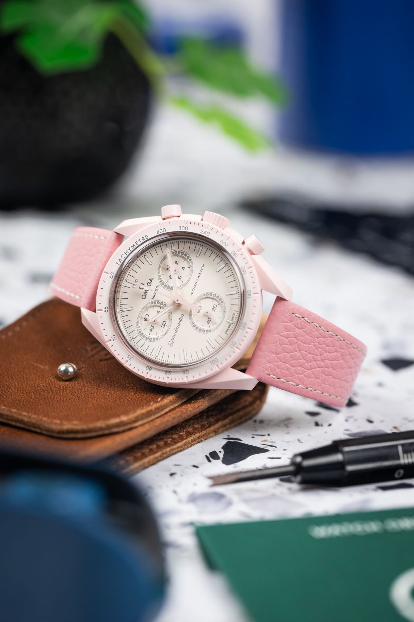 Close-up of a pink Omega chronograph wristwatch with a white dial and three subdials, mounted on a pink leather strap with white stitching, resting on a brown leather wallet. The watch is surrounded by a blurred workspace setting including a precision tool, green card, and soft-focus background elements.