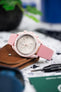 Close-up of a pink Omega chronograph wristwatch with a white dial and three subdials, mounted on a pink leather strap with white stitching, resting on a brown leather wallet. The watch is surrounded by a blurred workspace setting including a precision tool, green card, and soft-focus background elements.