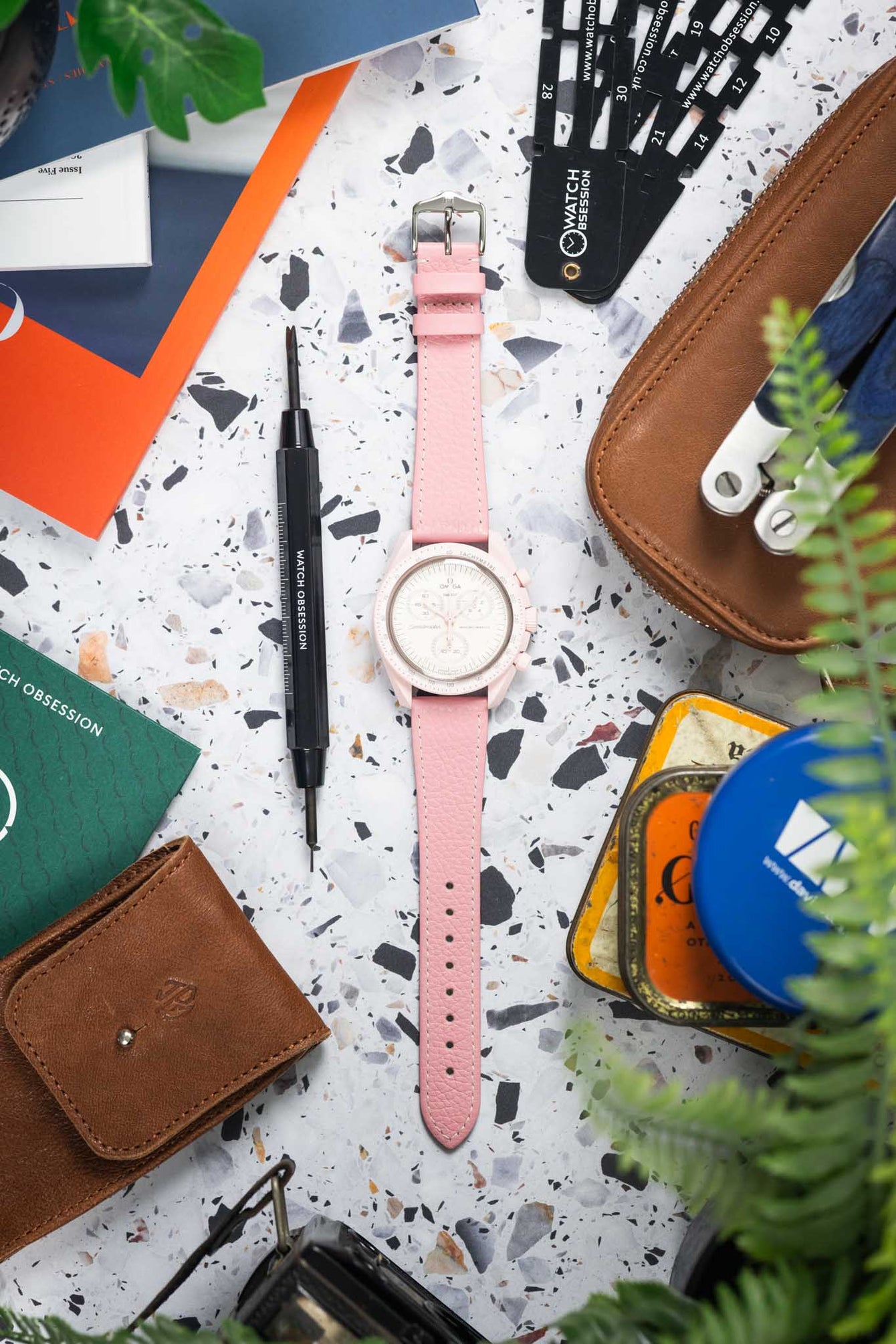 Top-down view of a pink chronograph wristwatch with a white dial and subdials, laid flat on a speckled surface and paired with a pink leather strap with white stitching. The watch is surrounded by everyday items including a brown leather wallet, a precision screwdriver, strap measuring tools labeled “Watch Obsession,” colorful cards, a tin container, and partially visible green plant leaves.