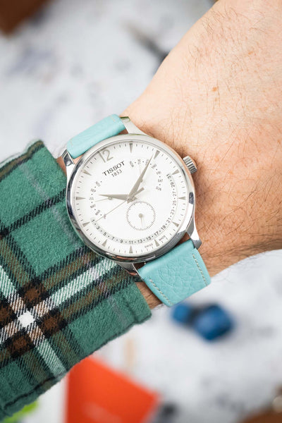 Close-up of a silver Tissot wristwatch with a white dial featuring day, date, and month indicators, worn on a wrist with a turquoise leather strap; a green plaid sleeve is visible, with a softly blurred, colorful background.