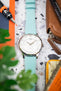 Top-down view of a silver Tissot wristwatch with a white dial and turquoise leather strap, placed on a speckled surface surrounded by everyday items including a brown leather pouch, a precision tool, colorful objects, and partially blurred green leaves in the foreground.
