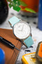 Silver Tissot wristwatch with a white dial and calendar display, paired with a turquoise leather strap, resting on a brown leather wallet with colorful blurred objects in the background.