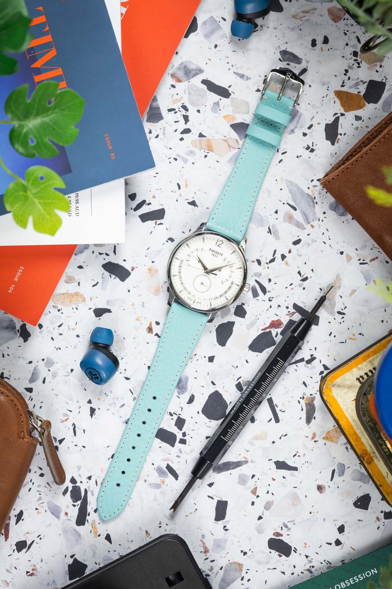Top-down view of a silver Tissot wristwatch with a white dial and turquoise leather strap laid flat on a speckled surface, surrounded by everyday items including a brown leather pouch, a precision screwdriver, wireless earbuds, colorful cards, and partially visible green plant leaves.