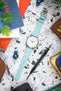 Top-down view of a silver Tissot wristwatch with a white dial and turquoise leather strap laid flat on a speckled surface, surrounded by everyday items including a brown leather pouch, a precision screwdriver, wireless earbuds, colorful cards, and partially visible green plant leaves.