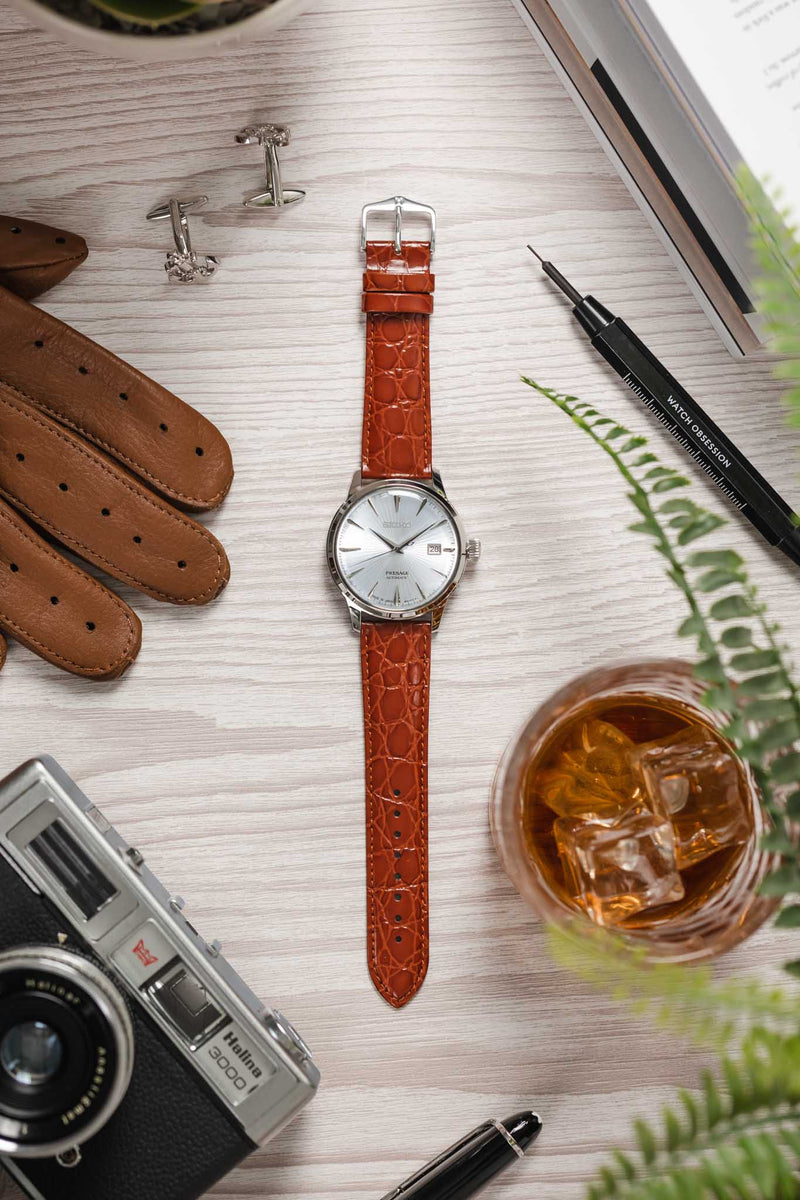 Flat lay of a Seiko dress watch with a silver dial on a gold‑brown crocodile-pattern strap, arranged on a wooden surface with a camera, gloves, cufflinks, a drink, a pen, and watch tools.