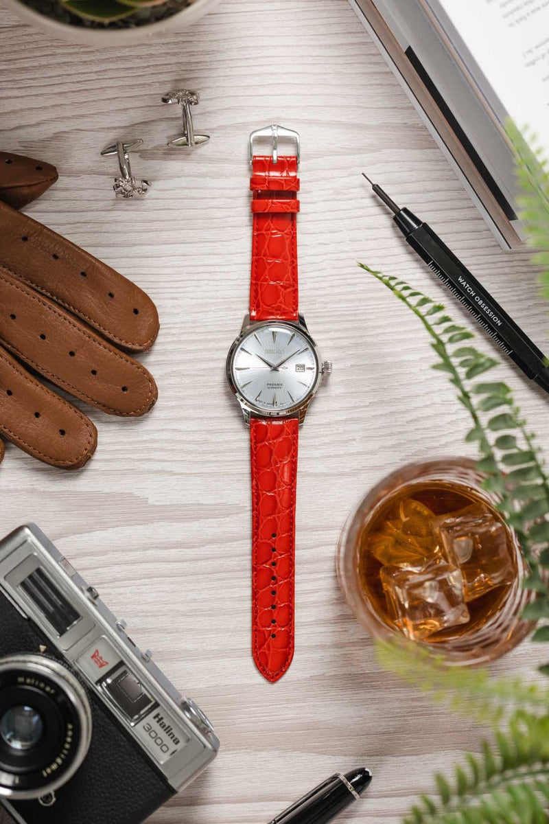 Flat lay of a Seiko dress watch with a white dial on a red crocodile-pattern strap, arranged on a wooden surface with gloves, cufflinks, a camera, a drink, tools, and greenery.