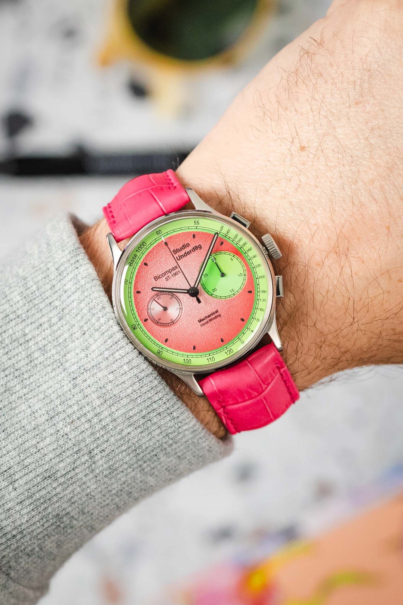 An overhead shot of a wrist adorned with a Studio Underd0g Bicompax Chronograph Watermelon watch, featuring a silver-toned case, a two-tone pink and green dial with sub-dials, and a vibrant hot pink Hirsch Duke leather strap with crocodile-style texture. The cuff of a light grey knit sweater is visible on the arm, and the background is blurred.