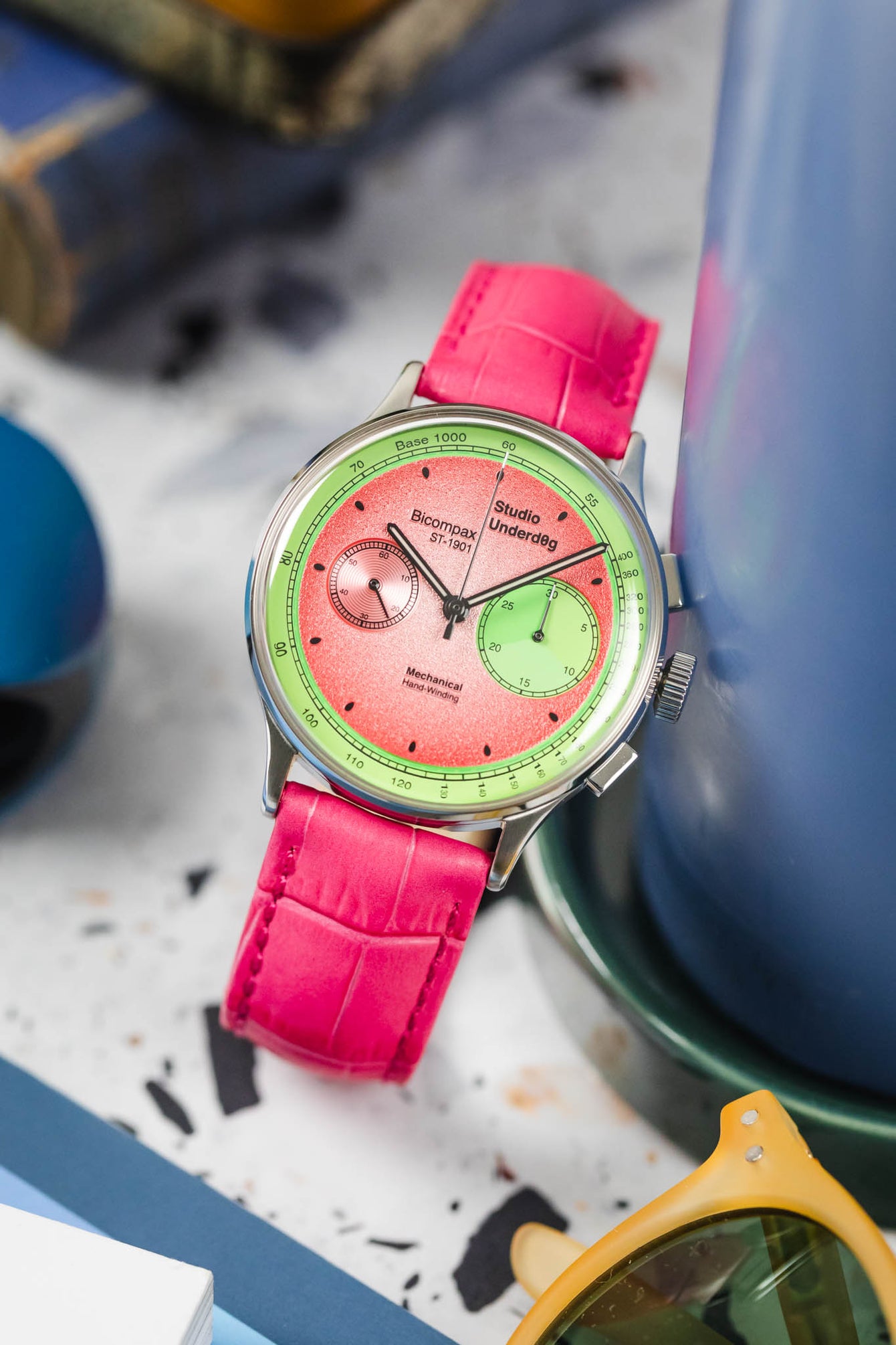 A Studio Underd0g Bicompax Chronograph Watermelon watch with a round stainless steel case and a unique pink and green dial with white sub-dials, is fitted with a hot pink Hirsch Duke leather strap with a crocodile-style texture. The watch is positioned at an angle on a light grey terrazzo-patterned surface. A tall blue cylindrical object is in the mid-ground, and yellow-tinted sunglasses are visible in the foreground.