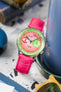 A Studio Underd0g Bicompax Chronograph Watermelon watch with a round stainless steel case and a unique pink and green dial with white sub-dials, is fitted with a hot pink Hirsch Duke leather strap with a crocodile-style texture. The watch is positioned at an angle on a light grey terrazzo-patterned surface. A tall blue cylindrical object is in the mid-ground, and yellow-tinted sunglasses are visible in the foreground.
