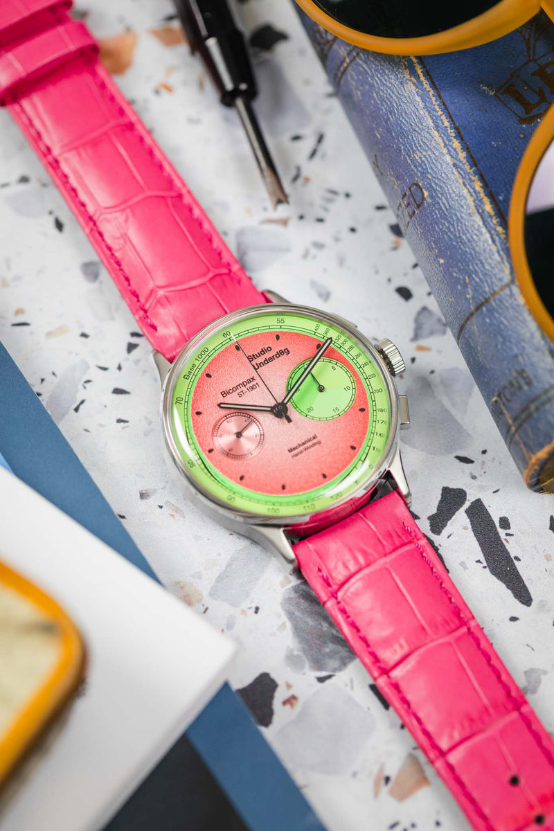 An eye-level close-up of a Studio Underdog Bicompax Chronograph Watermelon watch, showcasing its unique pink and green dial with white sub-dials and indices. The watch is fitted with a hot pink Hirsch Duke leather strap with crocodile-style embossing and a silver-toned tang buckle. The background is blurred, with hints of a blue object and yellow sunglasses.