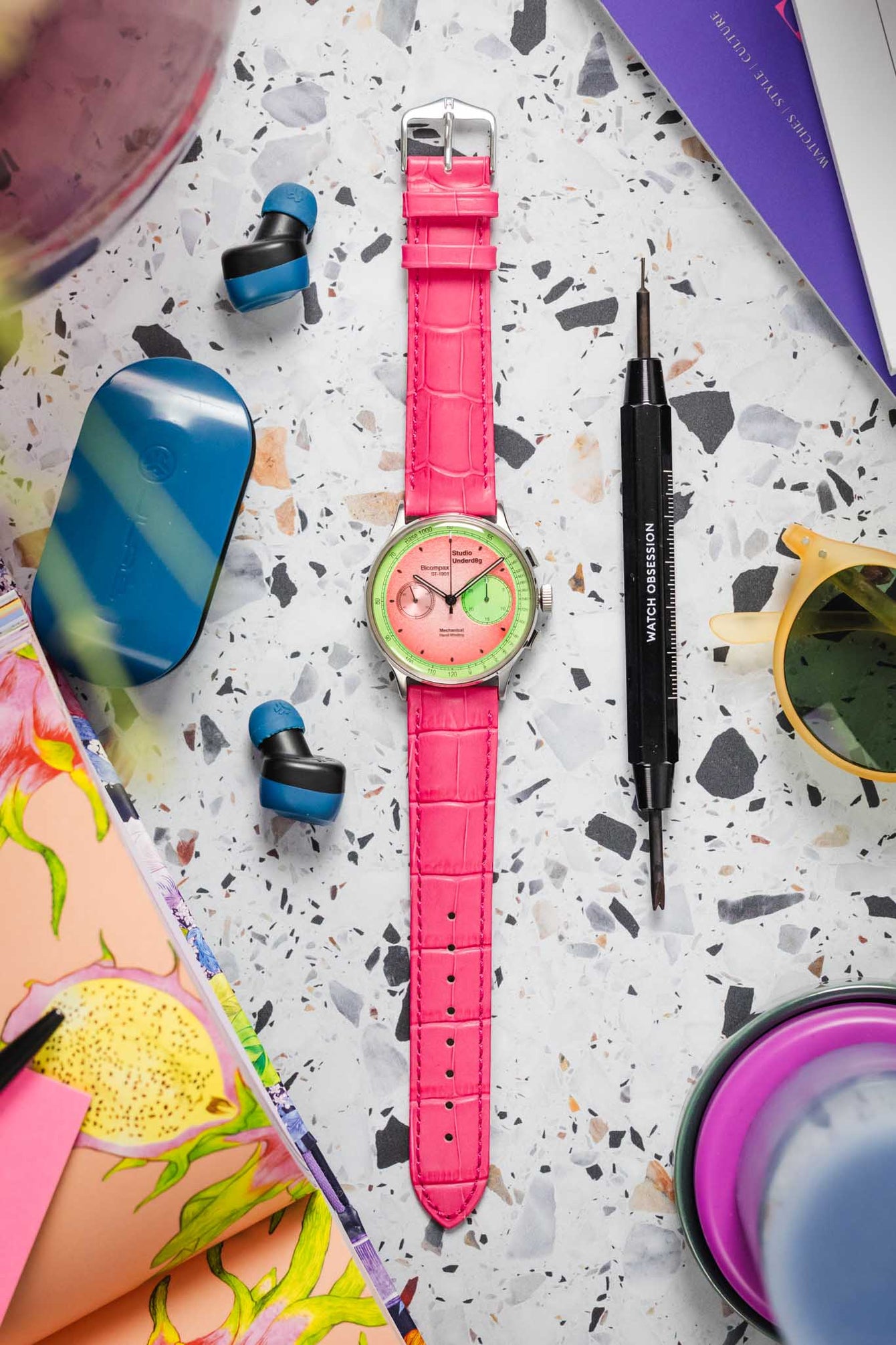 A vibrant chronograph watch, featuring a light pink and lime green two-tone dial with multiple sub-dials, is fitted with a hot pink Hirsch Duke leather strap with crocodile-style embossing. The watch is positioned diagonally on a light grey terrazzo-patterned surface. Surrounding it are blue wireless earbuds, a blue case, a black "WATCH OBSESSION" screwdriver, yellow-tinted sunglasses, and colorful books or magazines.