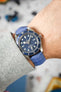 An overhead shot of a wrist adorned with a Tudor Black Bay 58 watch, featuring a silver-toned case, a blue dial with white indices and snowflake hands, and a light blue Hirsch Duke leather strap with crocodile-style texture. The cuff of a light grey knit sweater is visible on the arm.