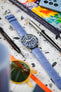  A Tudor Black Bay 58 watch, featuring a blue dial, blue rotating bezel, and a silver-toned case, is fitted with a light blue Hirsch Duke leather strap with crocodile-style embossing. The watch is positioned diagonally on a light grey terrazzo-patterned surface. Surrounding it are a black "WATCH OBSESSION" screwdriver, a silver pen, a vibrant book or magazine with abstract art, and blurred orange-tinted sunglasses in the foreground.