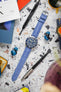 A Tudor Black Bay 58 watch with a blue dial and bezel, fitted with a light blue Hirsch Duke leather strap, is centered on a grey terrazzo-patterned surface. Surrounding the watch are a silver pen, yellow-tinted sunglasses, blue wireless earbuds, a black "WATCH OBSESSION" screwdriver, and various magazines or books, including one titled "ORACLE".