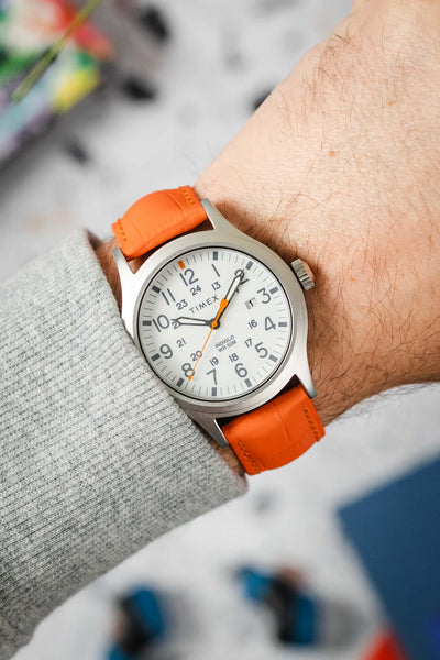A close-up of a person's wrist wearing a silver-toned Timex Expedition North Sierra watch with a white dial and an orange Hirsch Duke leather watch strap. The cuff of a grey sweater is visible.