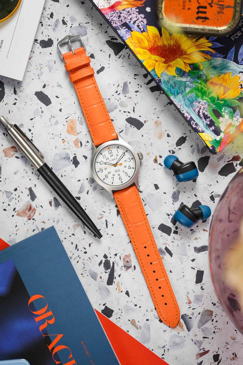 A detailed shot of a bright orange Hirsch Duke watch strap, highlighting its textured surface and polished silver-toned buckle. The strap is slightly curved, suggesting it is being worn.