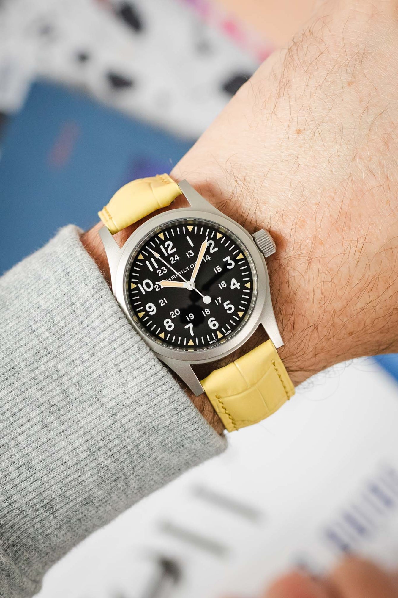  A Hamilton Khaki Field Mechanical watch, featuring a black dial with white Arabic numerals and a 24-hour scale, is fitted with a vibrant yellow Hirsch Duke leather watch strap that has a crocodile-style texture. The watch is positioned on a deep blue book or magazine, with a black pen in the foreground and blurred colored elements in the background.