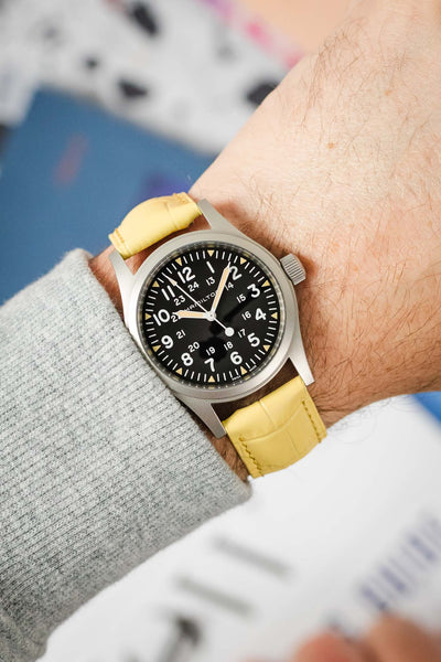  A Hamilton Khaki Field Mechanical watch, featuring a black dial with white Arabic numerals and a 24-hour scale, is fitted with a vibrant yellow Hirsch Duke leather watch strap that has a crocodile-style texture. The watch is positioned on a deep blue book or magazine, with a black pen in the foreground and blurred colored elements in the background.
